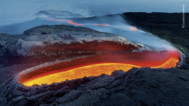 Etna's river of fire by Luciano Gaudenzio, Italy. Winner 2020, Earth's Environments. From a great gash on the southern flank of Mount Etna, lava flows within a huge lava tunnel, re-emerging further down the slope as an incandescent red river, veiled in volcanic gases. To witness the scene, Luciano and his colleagues had trekked for several hours up the north side of the volcano, through stinking steam and over ash-covered chaotic rocky masses &ndash; the residues of past eruptions. A wall of heat marked the limit of their approach. Luciano describes the show that lay before him as hypnotic, the vent resembling &lsquo;an open wound on the rough and wrinkled skin of a huge dinosaur&rsquo;. It was 2017, and he had been on the nearby island of Stromboli to photograph eruptions there when he heard news of the new vent on what is Europe&rsquo;s largest volcano. He took the very next ferry, hoping he would arrive in time to see the peak of the latest show.