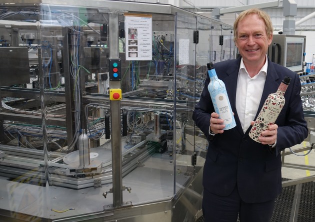 Frugalpac CEO Malcolm Waugh holds Mothers Ruined Gin and Cantina Goccia Frugal Bottles alongside the company's Frugal Bottle Assembly Machine.