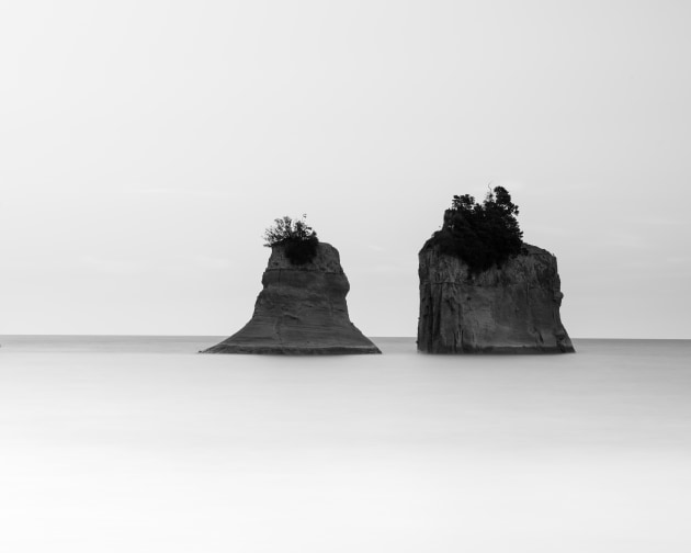 Taranaki coastline, New Zealand. Two lone rock stacks sit isolated by a long exposure that smooths the sea to a silky plane, the twin forms floating in quiet solitude against a minimal sky. Nikon D810, Nikkor 70–200mm f4 lens @ 112mm, 30s @ f11, ISO 100.