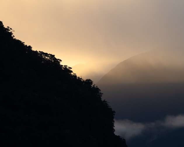 Fiordland, New Zealand. Light breaks through the mist as it slips toward the horizon, adding contrast between the receding ridgelines falling into shadow and the warm glow beyond, naturally simplifying this complex landscape into bold, graphic shapes. Nikon D850, Nikkor 70–200mm f4 lens @ 200mm, 1/320s @ f8, ISO 400.